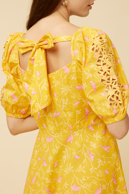 Close-up of the intricate cutwork detailing on the back and sleeves of the Peela Sunshine Dress, set against a backdrop of yellow fabric with leaf and bird motifs